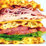 Picture of a Club Sandwich Chaffle cut in half on a white plate. Loaded with turkey, ham, cheese and bacon.