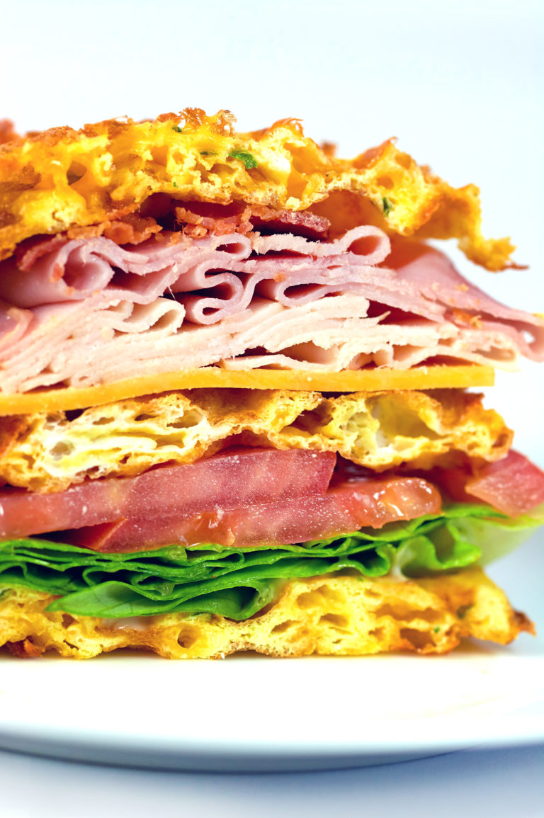 Picture of a Club Sandwich Chaffle cut in half on a white plate. Loaded with turkey, ham, cheese and bacon.