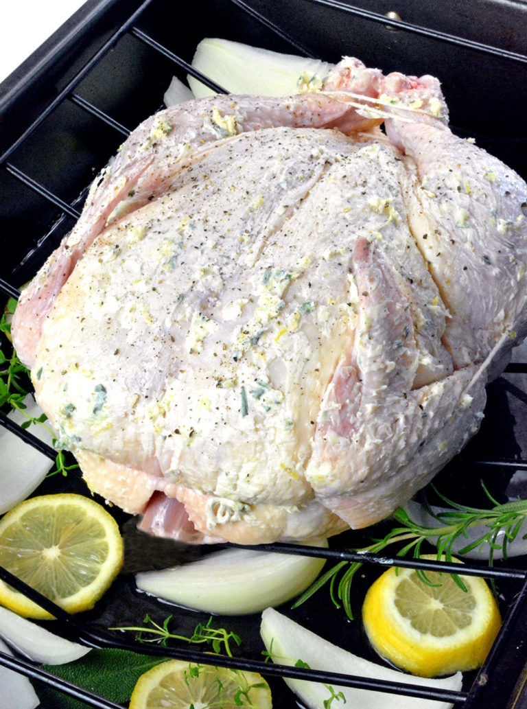 Raw whole chicken in a black roasting pan surrounded by lemon, onion and herbs.