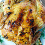 Whole chicken on a white plate surrounded by lemon, onion and herbs.