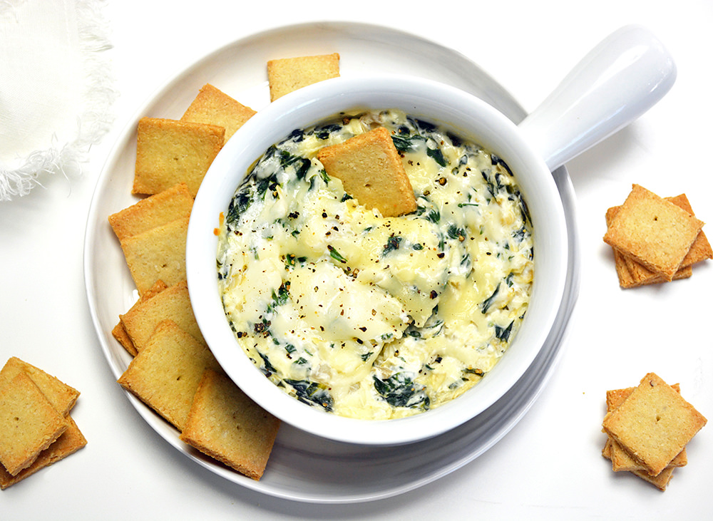 Baked Keto Spinach Dip in a white soup bowl. Rests on a white plate with keto crackers.