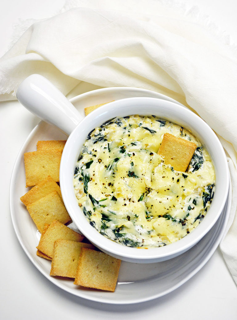 Baked Keto Spinach Dip in a white soup bowl. Rests on a white plate with keto crackers.