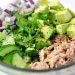 Clear glass bowl containing ingredients for tuna with avocado salad: tuna, cucumber, avocado, onions and cilantro