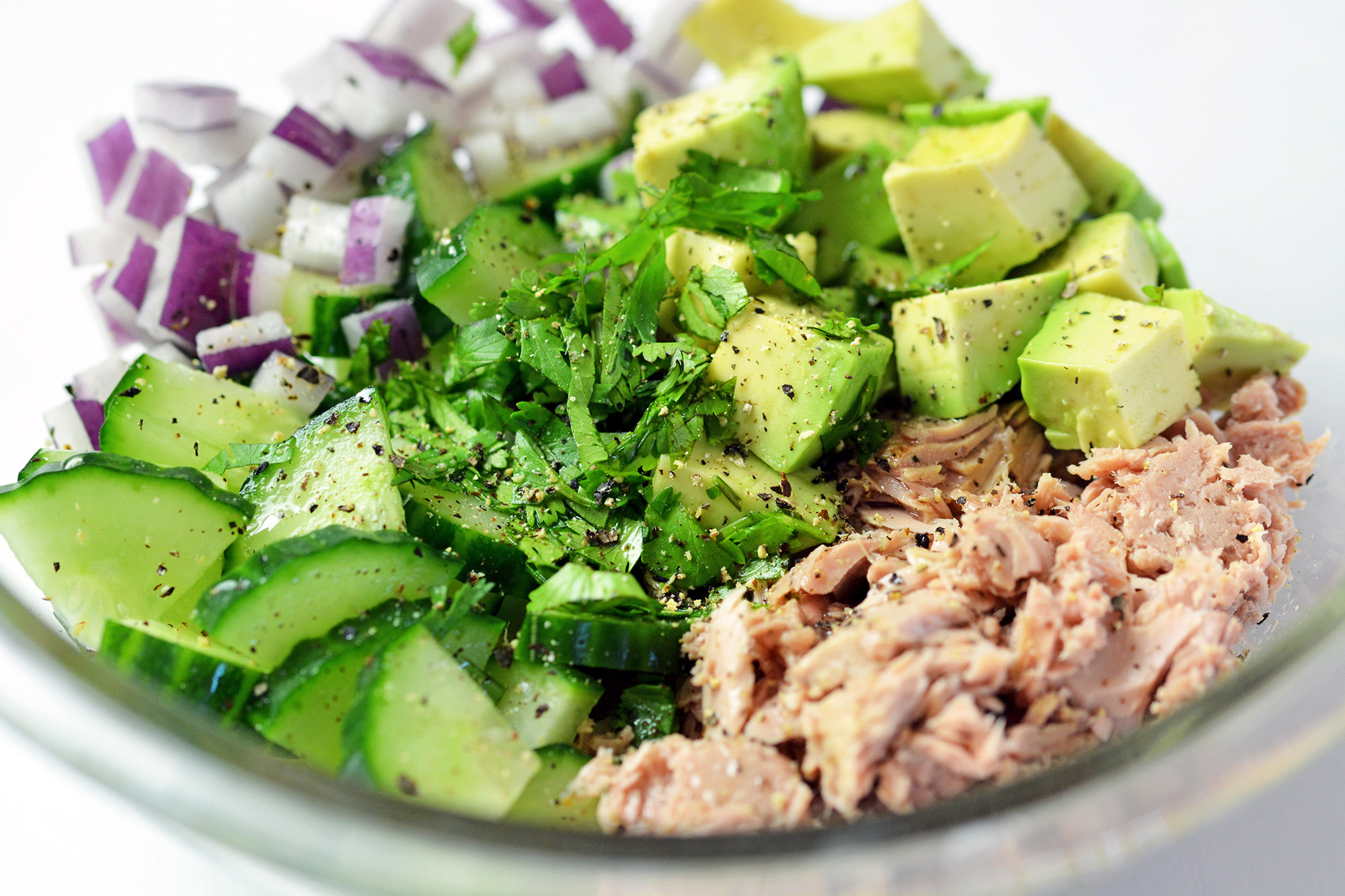 Clear glass bowl containing ingredients for tuna with avocado salad: tuna, cucumber, avocado, onions and cilantro