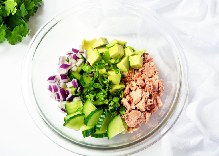 Clear glass bowl containing ingredients for tuna with avocado salad: tuna, cucumber, avocado, onions and cilantro