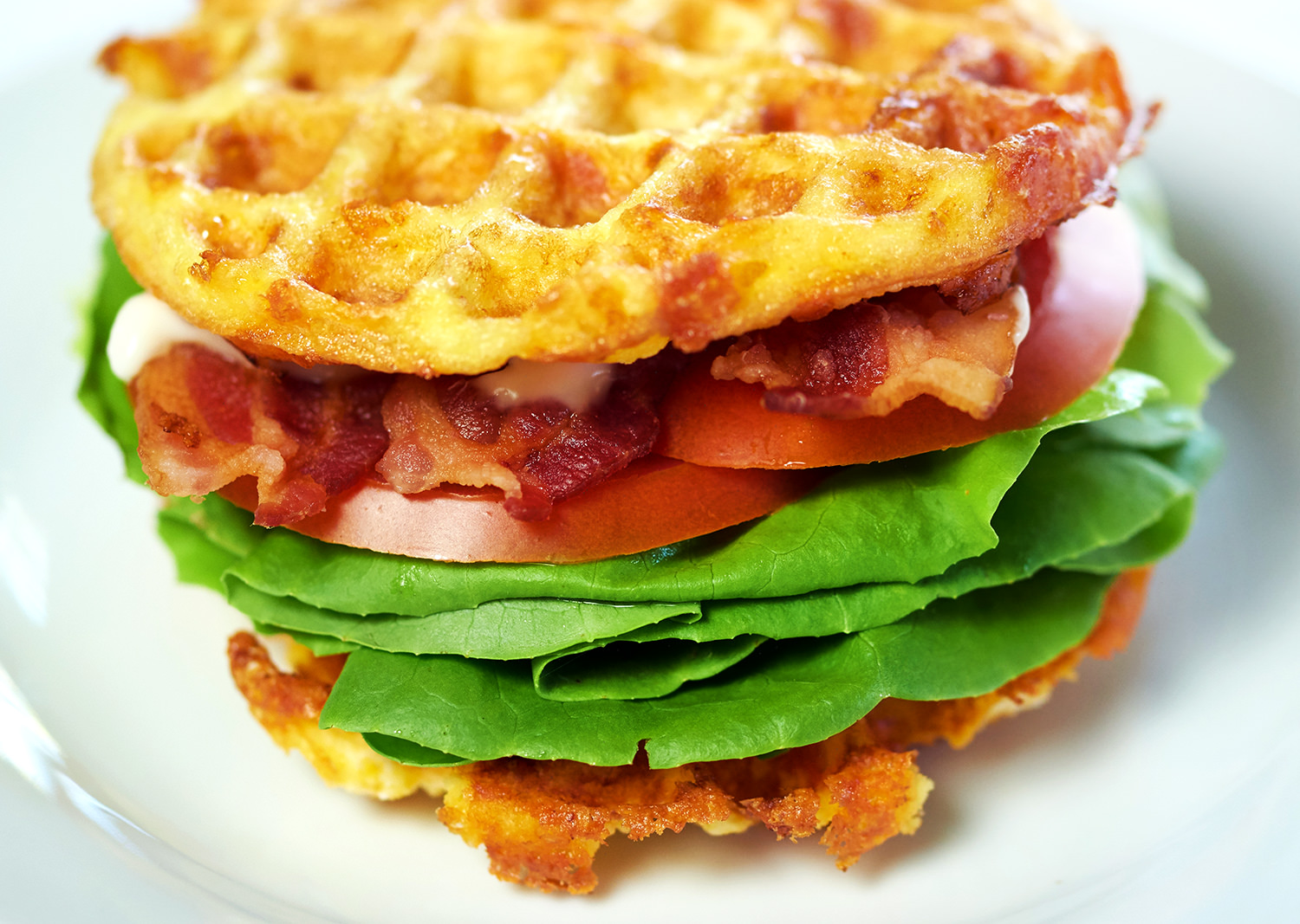 Picture of a BLT Chaffle Recipe on a white plate. The chaffle is loaded with lettuce, bacon, tomato and mayo.