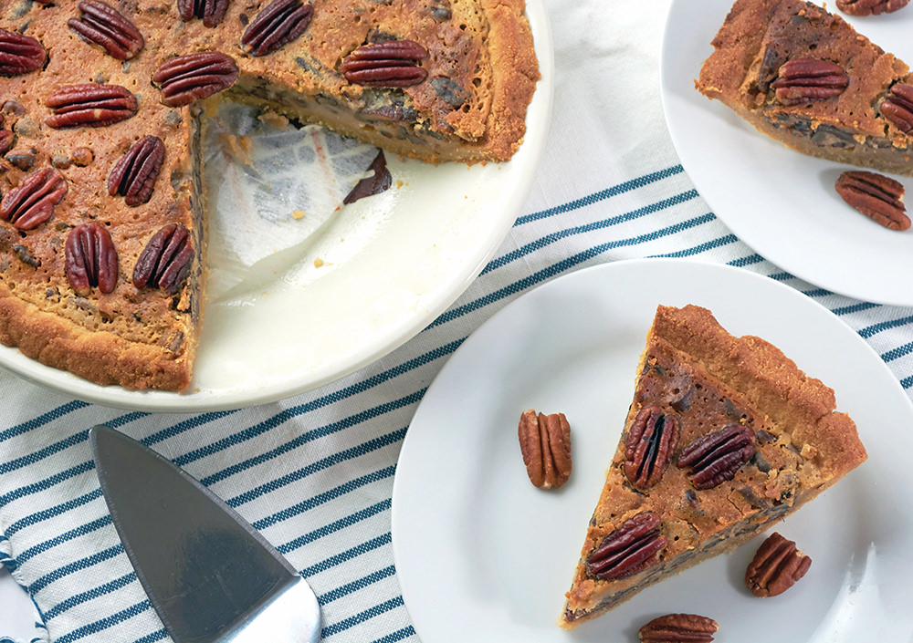 Whole keto pecan pie with two slices taken out and plated on a white plate and blue stripped napkin.