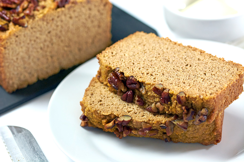 Two slices of Keto Pumpkin bread on a black cutting board. The remaining loaf is pictured in the background