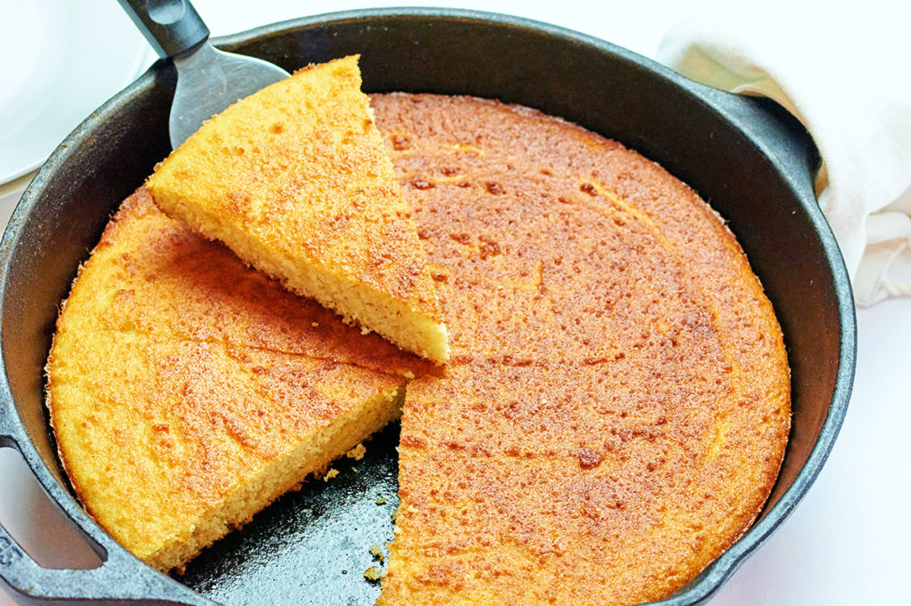 keto cornbread in a round black cast iron skillet. Once triangular slice is cut out and being held above the skillet on top of a serving utensil