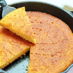 keto cornbread in a round black cast iron skillet. Once triangular slice is cut out and being held above the skillet on top of a serving utensil