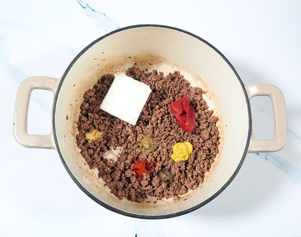 A cream colored dutch oven containing cooked ground beef, cream cheese, seasonings, ketchup and mustard sitting on top of the beef.