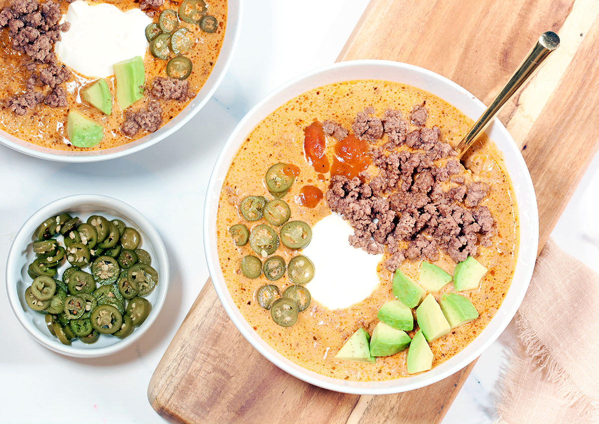 A white bowl of Keto Cheeseburger soup topped with beef, jalapenos, avocado and sour cream. A white bowl of jalapenos and a second bowl of soup is next to it.