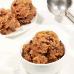 scoops of raw chocolate chip cookie dough in small white bowl. ice cream scooper in the back ground