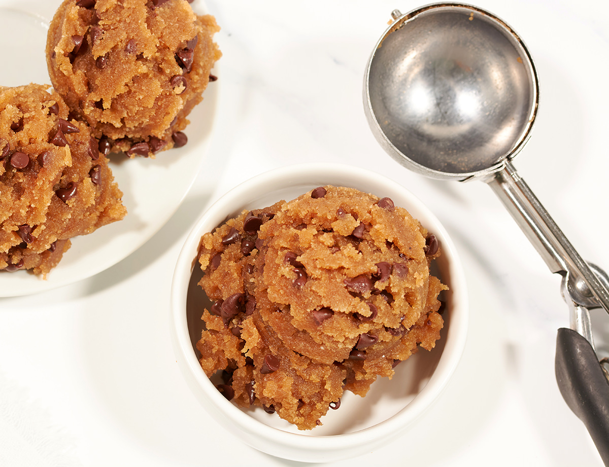 single scoop of raw cookie dough in small white bowl. Two scoops of raw cookie dough on small white plate. Cookie dough scooper off to side.