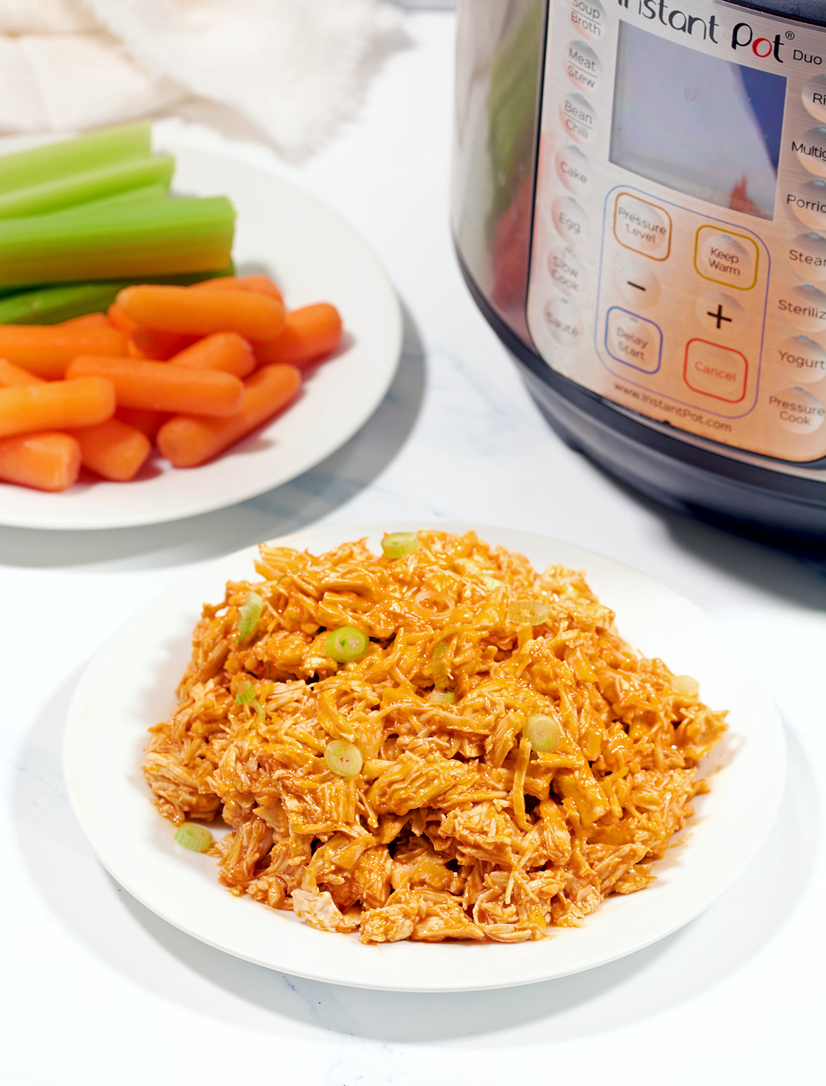 A bowl full of shredded buffalo chicken next to an Instant Pot and plate full of carrots and celery.