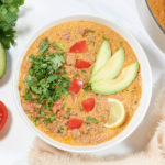 A white bowl of Keto Taco soup topped with cilantro, diced tomatoes, avocado and lime slice. A sliced tomatoes, bunch of cilantro, halved avocado and a pot of soup is next to it.