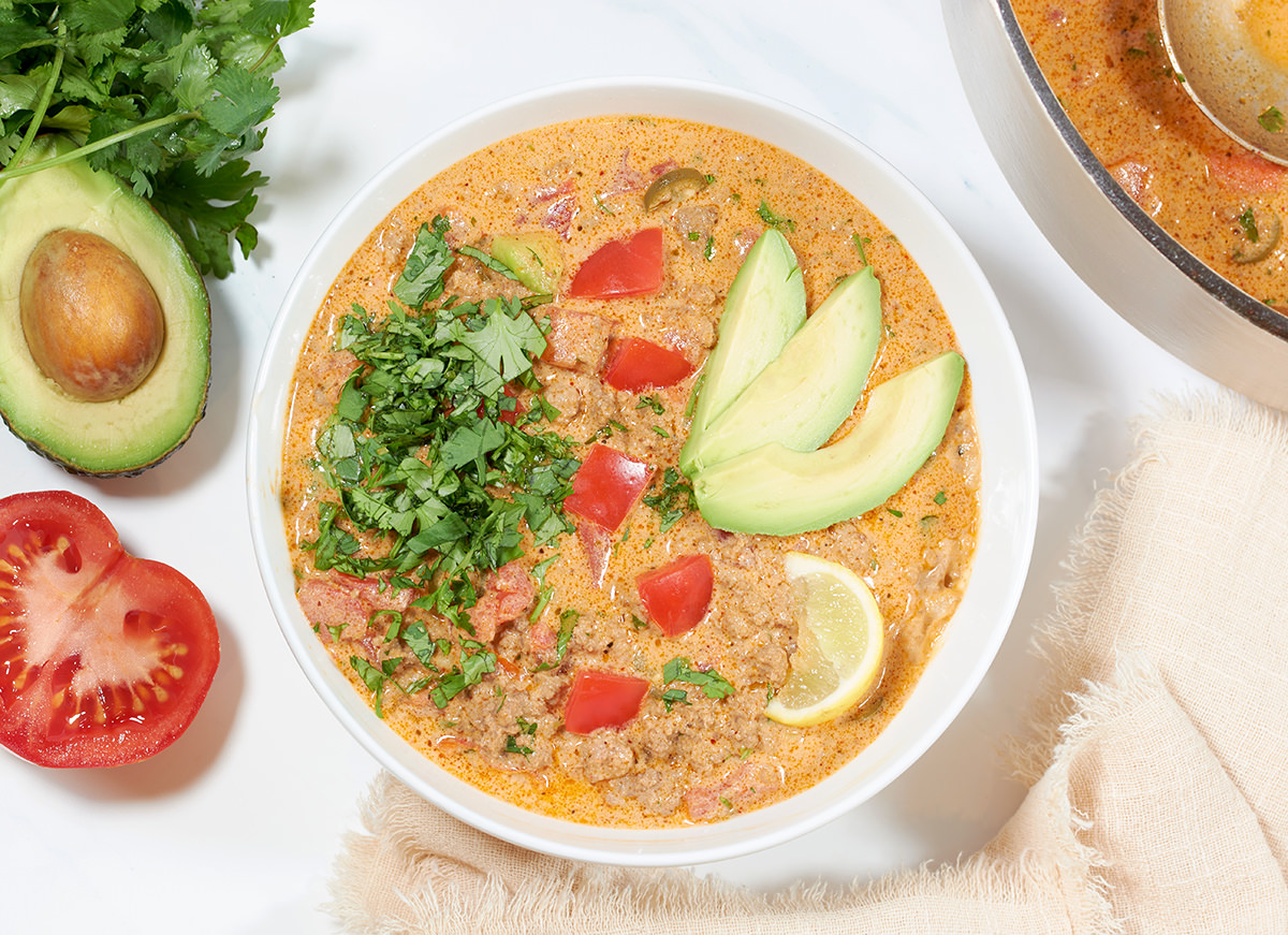 A white bowl of Keto Taco soup topped with cilantro, diced tomatoes, avocado and lime slice. A sliced tomatoes, bunch of cilantro, halved avocado and a pot of soup is next to it.