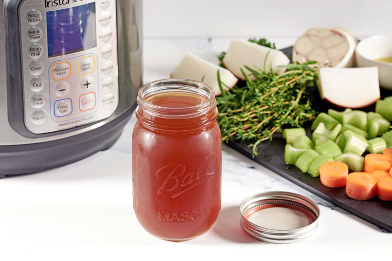 A mason jar full of beef broth in front of an instant pot. Cut veggies are on a black cutting board next to it.