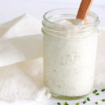 Ranch Dressing in glass mason jar with wooden spoon inside jar. Chopped chives are scattered around the jar. White cloth napkin is surrounding jar.