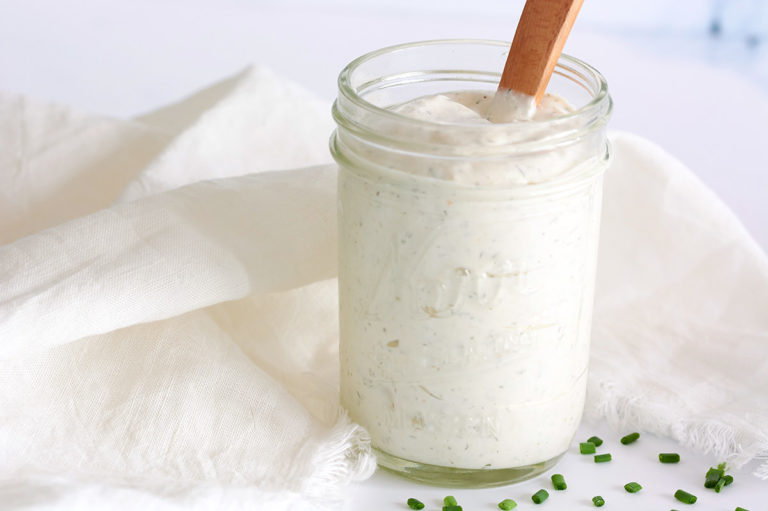 Ranch Dressing in glass mason jar with wooden spoon inside jar. Chopped chives are scattered around the jar. White cloth napkin is surrounding jar.