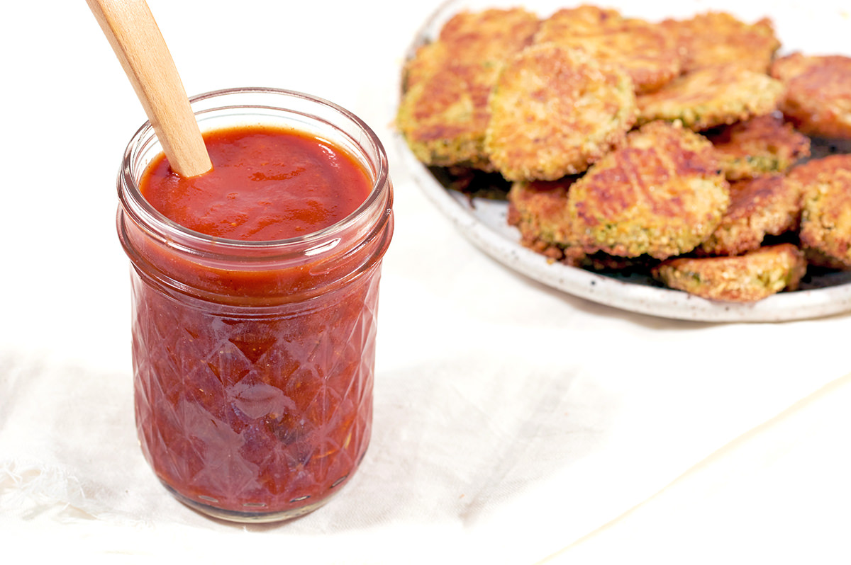 BBQ sauce in a glass mason jar with a wooden spoon inside. White Plate of fried pickles is in the background.