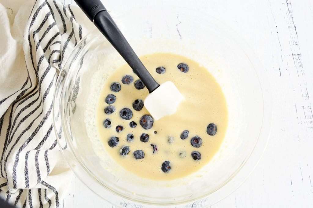 Clear bowl full of muffin mix and blueberries with a black spatula.