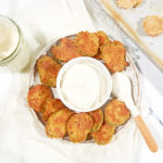 Keto fried pickles on a plate circling a small bowl of ranch dressing. Small wooden spoon, dipped in ranch, is sitting on the edge of the plate. Cookie sheet with uncooked, breaded pickles is off to the right side. Glass mason jar of ranch dip is to the left of the plate.