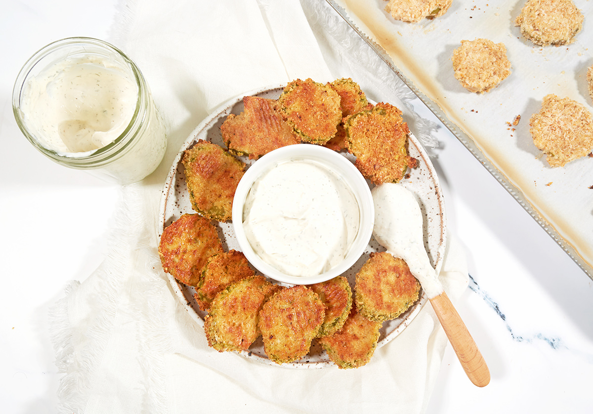 Keto fried pickles on a plate circling a small bowl of ranch dressing. Small wooden spoon, dipped in ranch, is sitting on the edge of the plate. Cookie sheet with uncooked, breaded pickles is off to the right side. Glass mason jar of ranch dip is to the left of the plate.