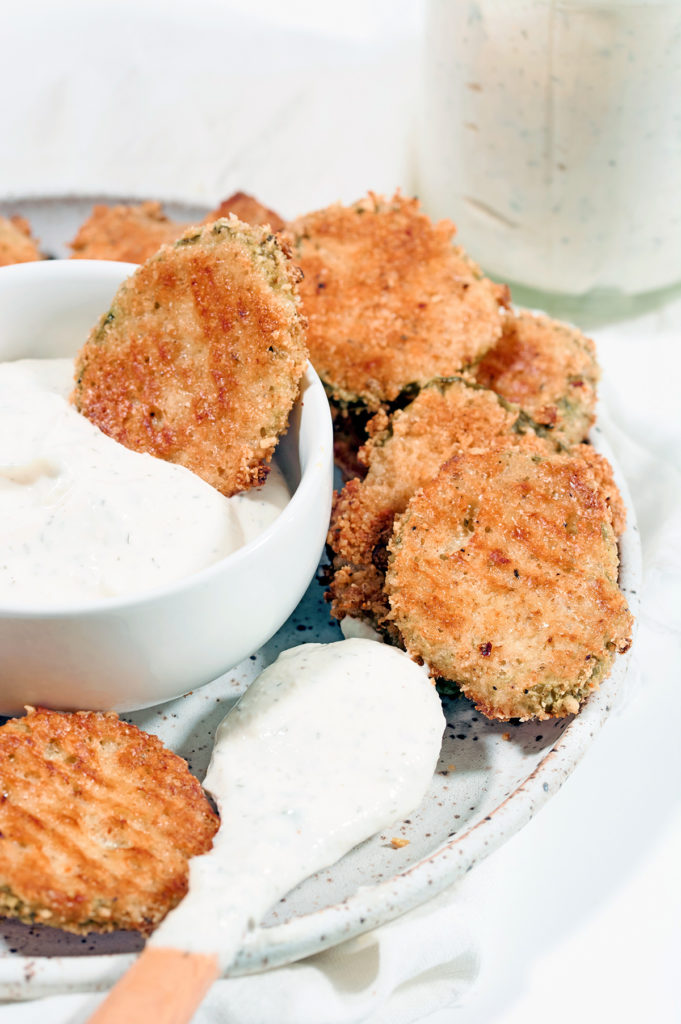 Pile of keto fried pickles on a white plate. Small white bowl of ranch in middle of bowl. One fried pickle is dipped in bowl of ranch. Wooden spoon dipped in ranch, is resting on plate.