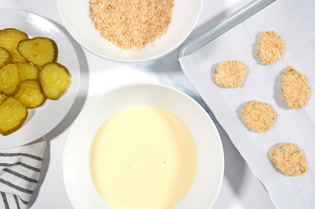 A large bowl of fried pickle breading next to a white bowl of unbreaded pickles and a silver baking sheet of breaded pickles.