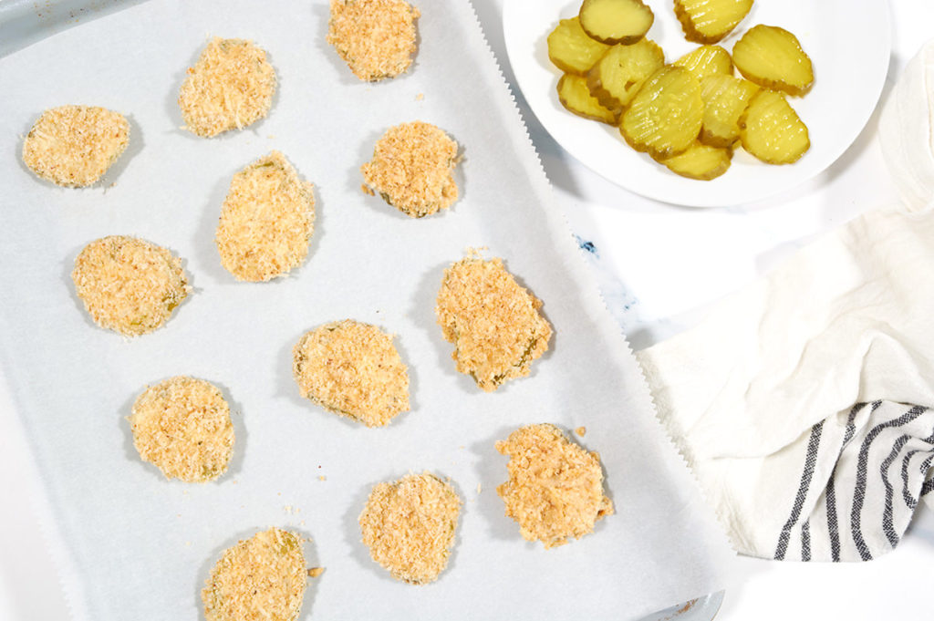 Uncooked breaded pickles on a silver baking tray next to a white napkin and plate of unbreaded pickles.