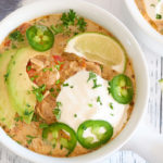 A white bowl filled with Keto white chicken chili. Chili is topped with avocado slices, slice of lime, sour cream, sliced fresh jalapeños & chopped up cilantro. Another bowl of chili is in the background.