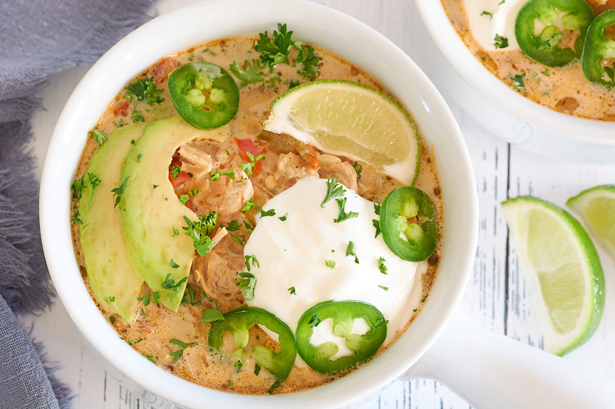 A white bowl filled with Keto white chicken chili. Chili is topped with avocado slices, slice of lime, sour cream, sliced fresh jalapeños & chopped up cilantro. Another bowl of chili is in the background.