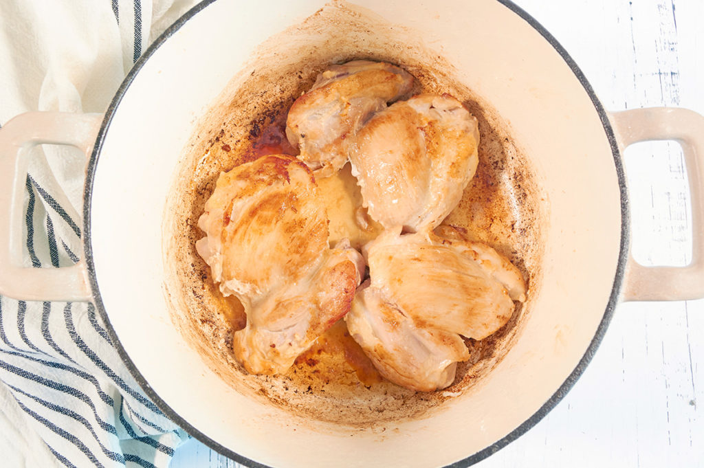 Browned chicken thighs in a cream dutch oven. The dutch oven is sitting on a white wood background next to a blue stripped kitchen towel.