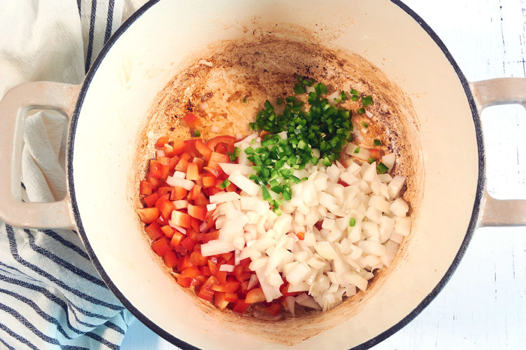 Chopped bell pepper, jalapeno and onions in a cream colored dutch oven. The dutch oven is sitting next to a blue stripped kitchen towel on a white wood counter.