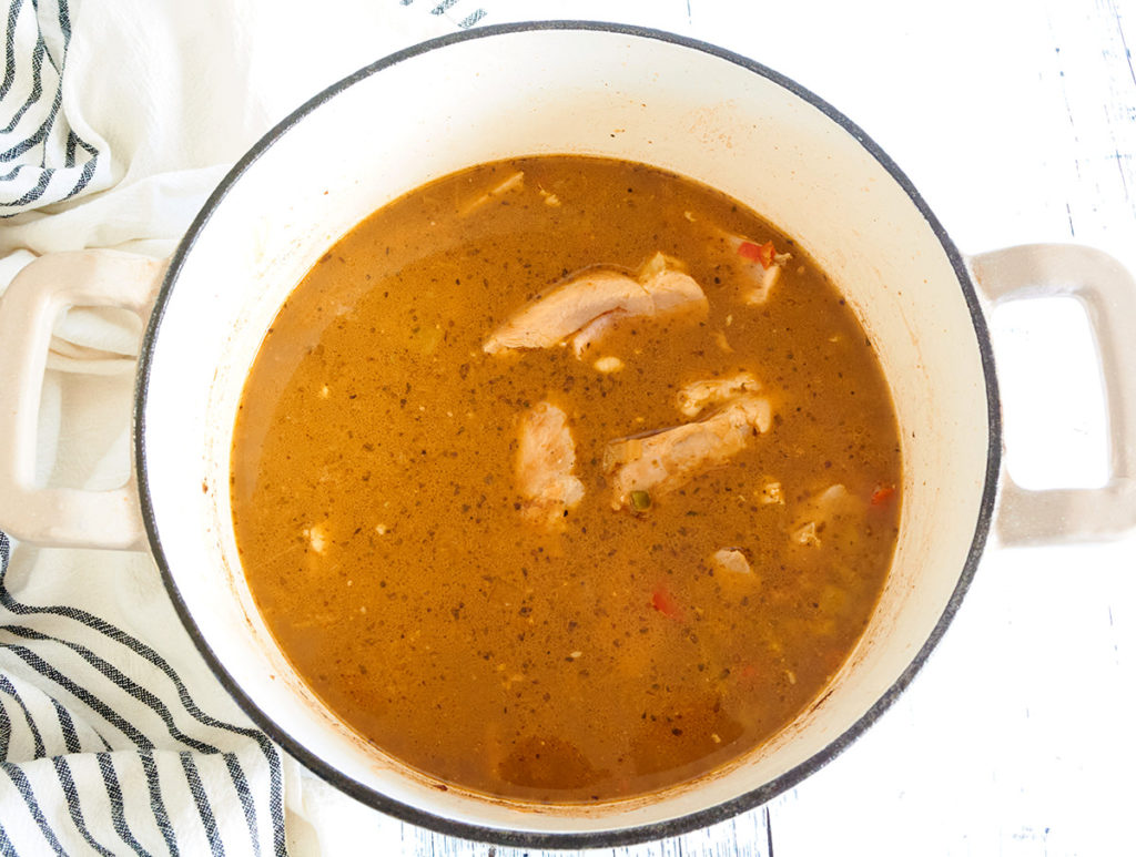 A cream dutch oven loaded with soup broth and chicken. It's sitting on a white background and blue striped kitchen towel.