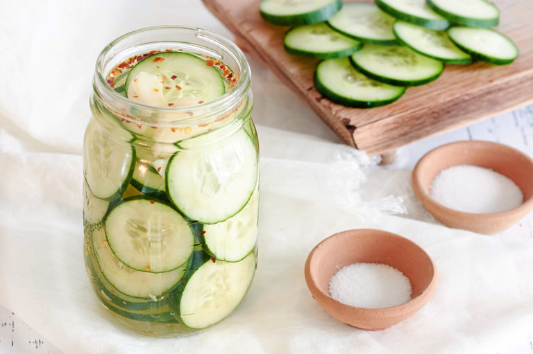 Jar of pickled cucumbers next to sliced cucumbers and salt and sugar.